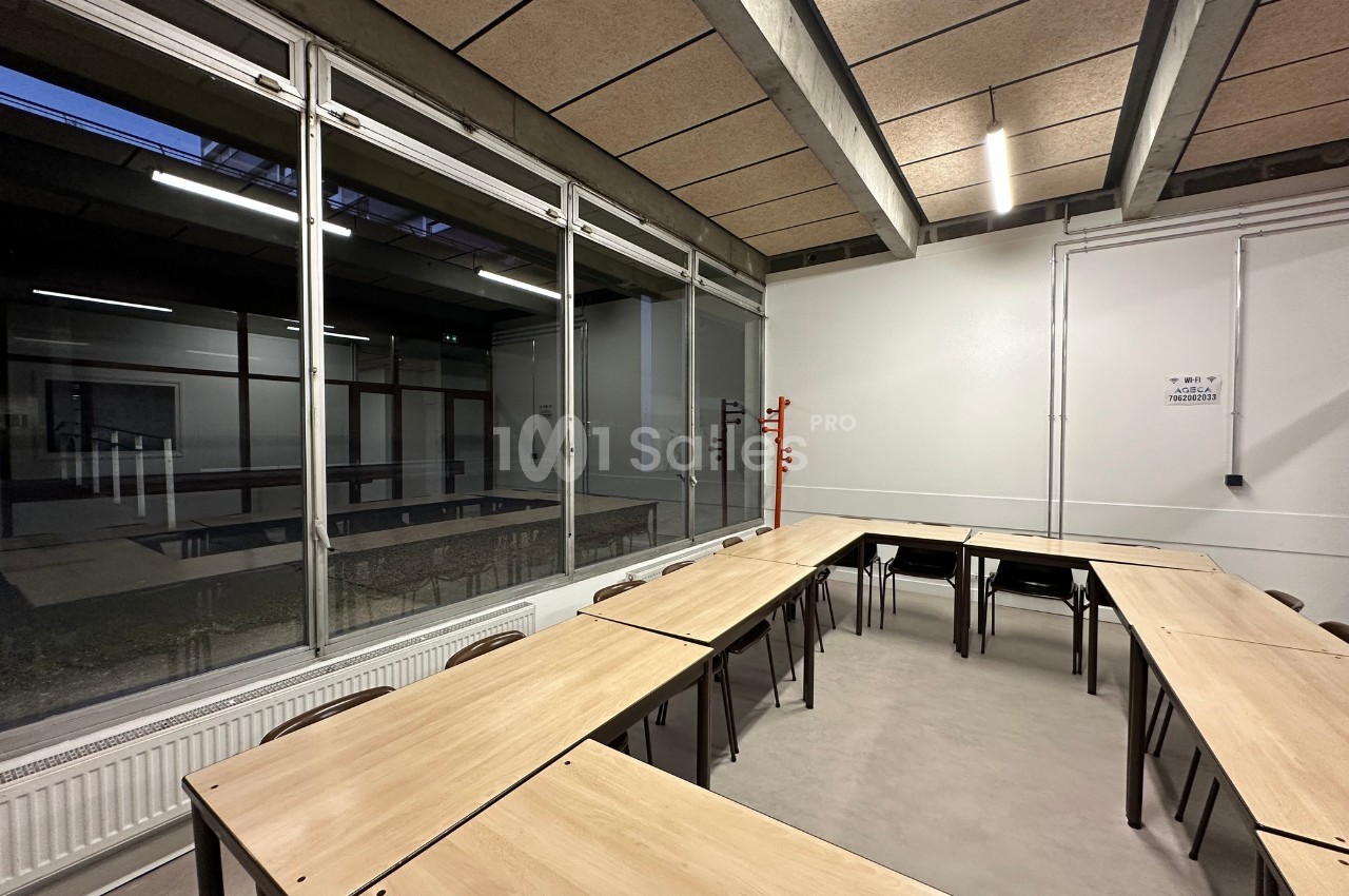 Salle de classe vide avec des tables disposées en U, éclairée par des néons et une grande baie vitrée donnant sur un couloir.