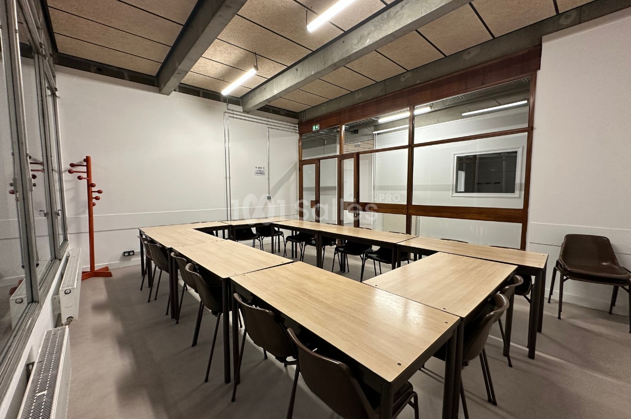 Salle de réunion avec tables disposées en U, chaises en plastique, murs blancs et grande baie vitrée.