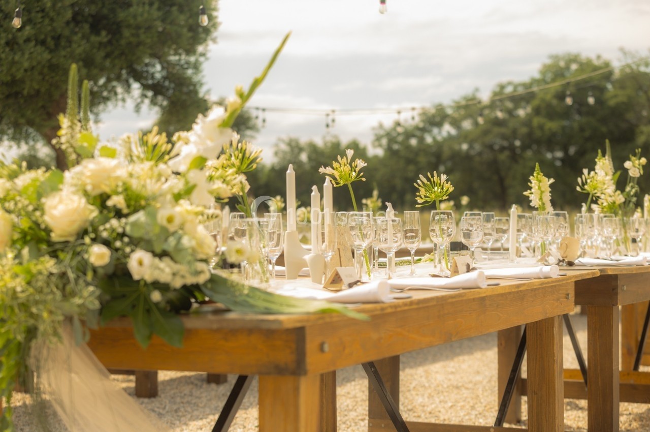 Table en bois décorée de fleurs blanches, bougies et vaisselle, installée en extérieur sous un ciel lumineux.