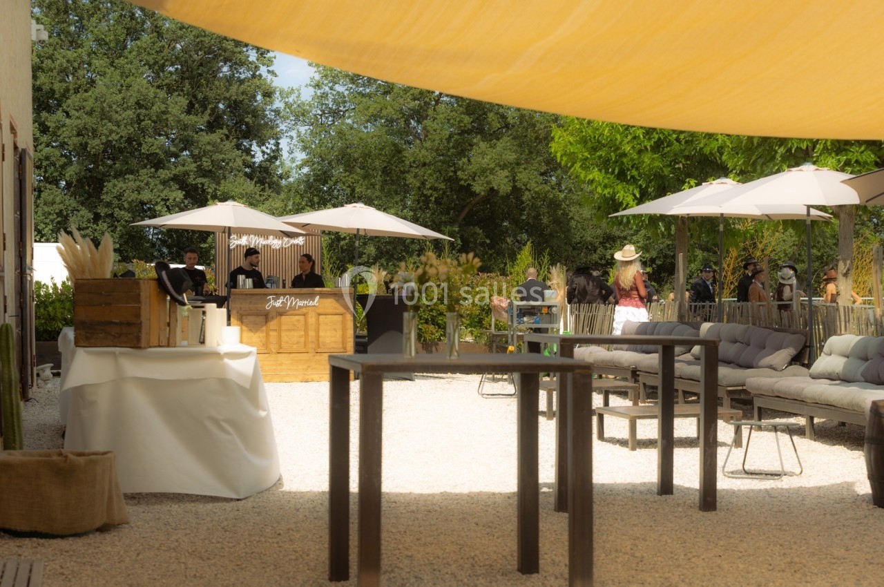 Terrasse extérieure ensoleillée avec tables hautes, canapés, parasols et un bar en arrière-plan.