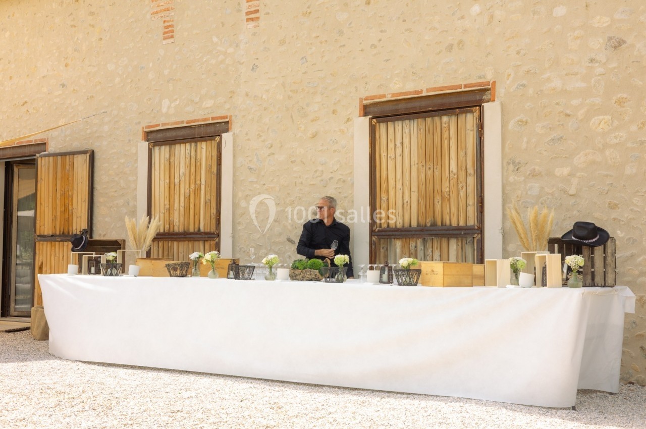 Homme debout derrière une longue table dressée avec des décorations florales, devant un mur en pierre beige.