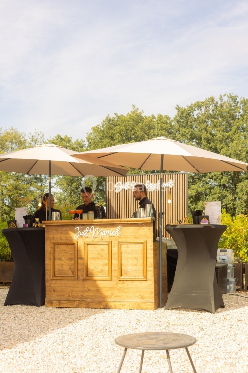 Stand en bois avec inscription ’Just Married’, abrité par des parasols, entouré de végétation et de ciel dégagé.