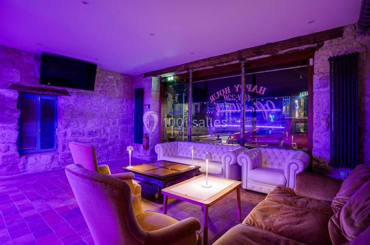 Salon cosy avec canapés et fauteuils éclairés par une lumière violette, grandes fenêtres donnant sur une rue urbaine.