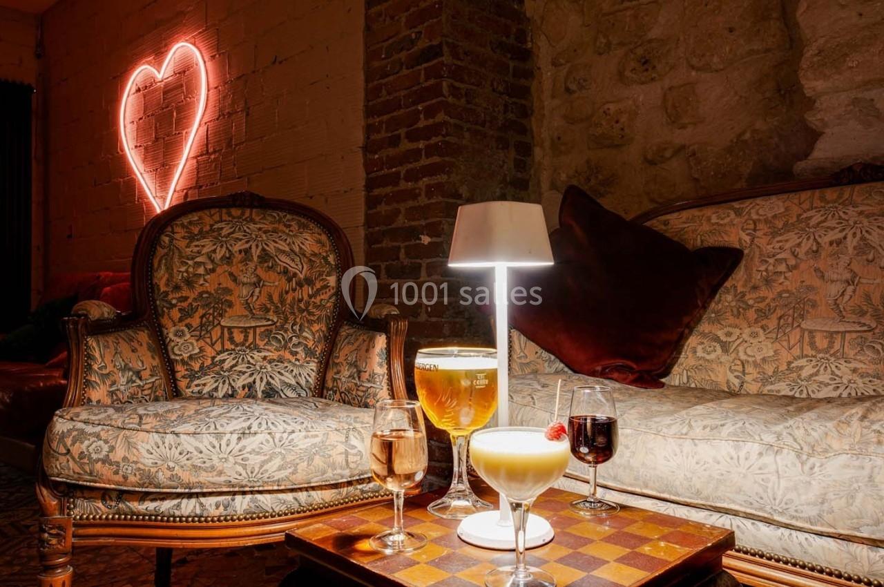 Deux fauteuils anciens entourent une table avec des cocktails, éclairée par une lampe, devant un mur avec un néon en forme…