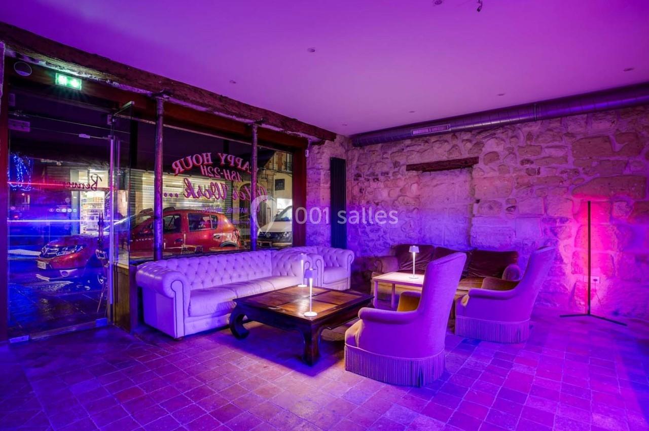 Salon avec canapé blanc, fauteuils et table basse dans une ambiance lumineuse violette, murs en pierre apparente.