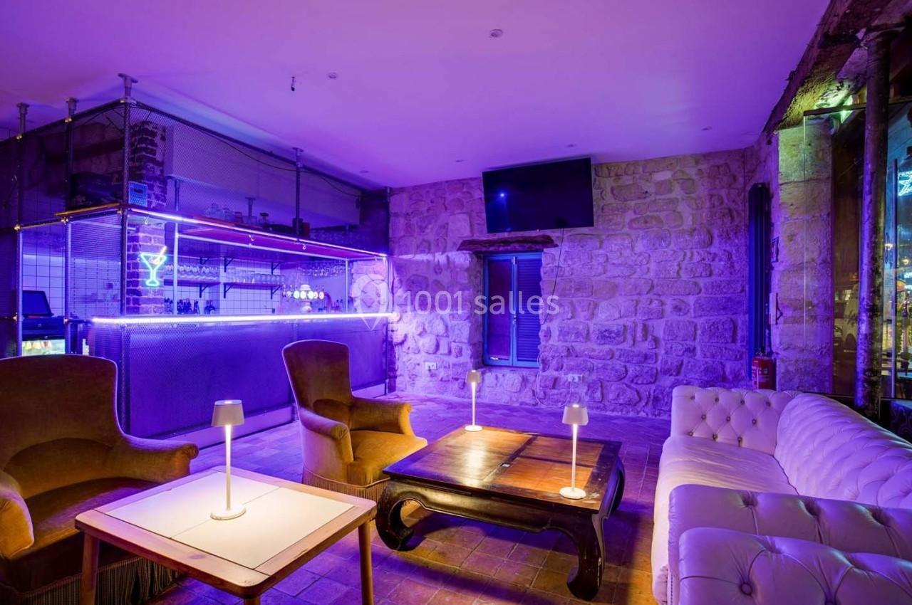 Salon avec bar éclairé par des néons violets, murs en pierre, fauteuils, canapé blanc et tables basses.