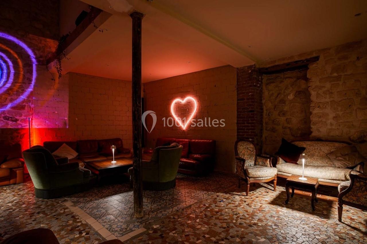 Salon cosy avec fauteuils, canapés, lumière tamisée et un néon en forme de cœur rouge sur un mur en pierre.