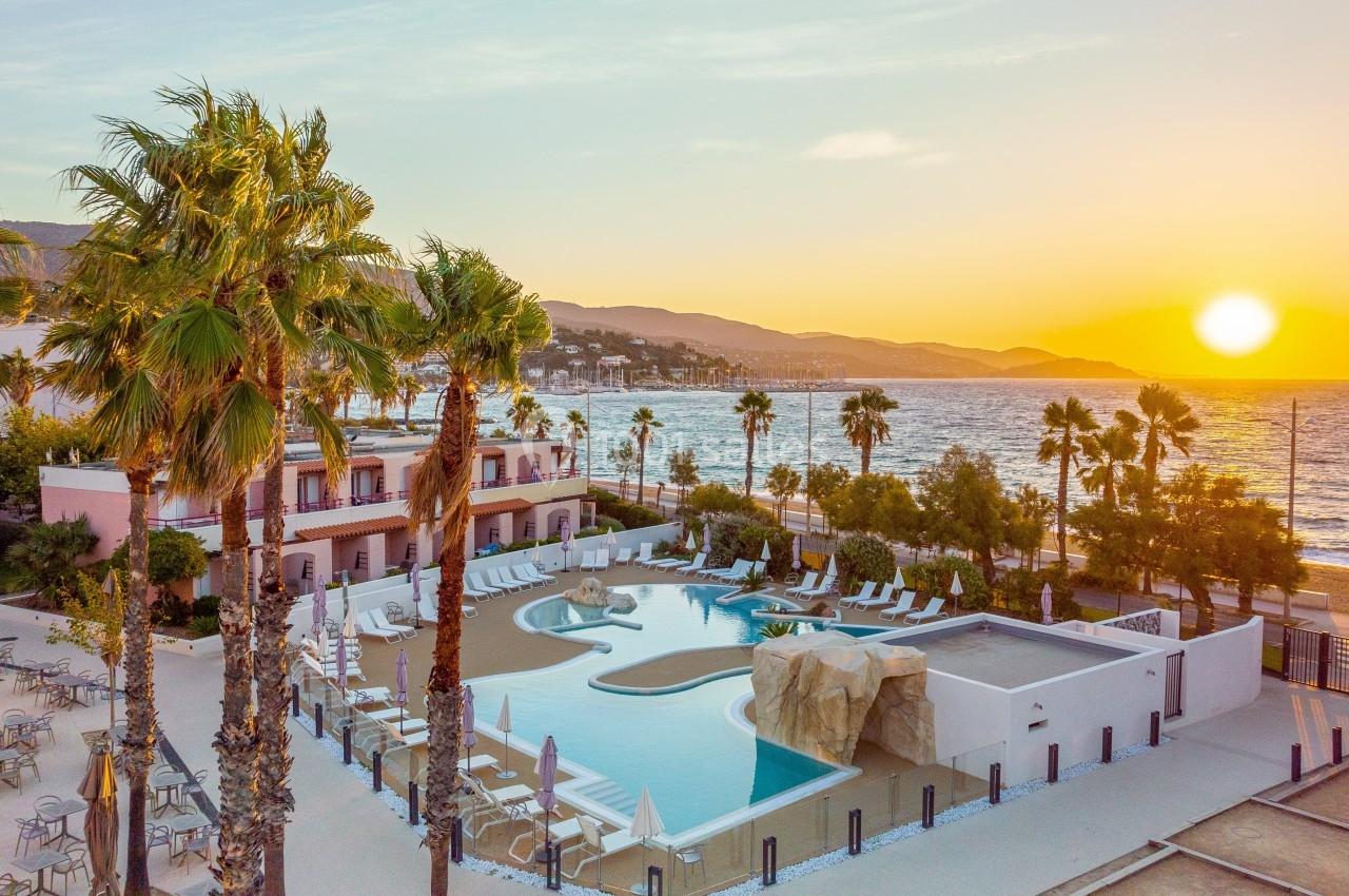 Vue d'une piscine extérieure bordée de palmiers, avec la mer et un coucher de soleil en arrière-plan.