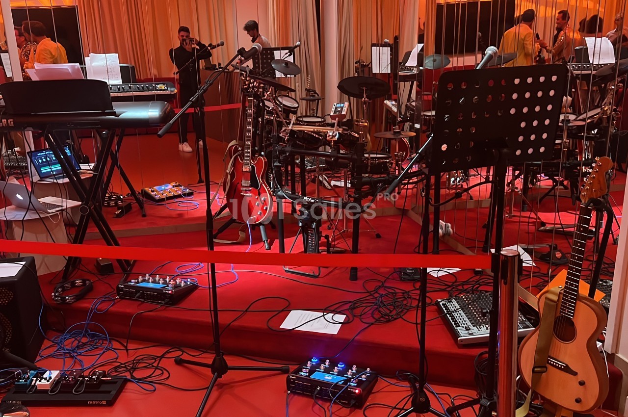 Salle de répétition avec instruments de musique, micros, câbles au sol et musiciens en arrière-plan.