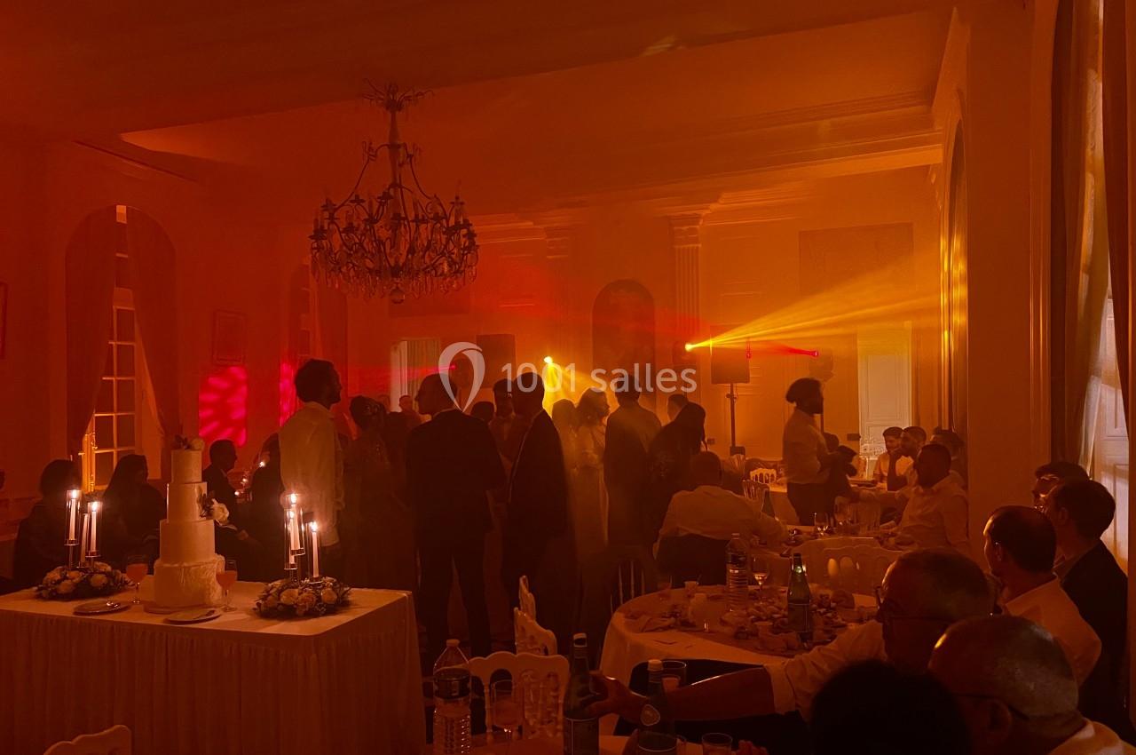 Salle de réception tamisée avec invités assis et debout, éclairage chaleureux et table avec gâteau de mariage.