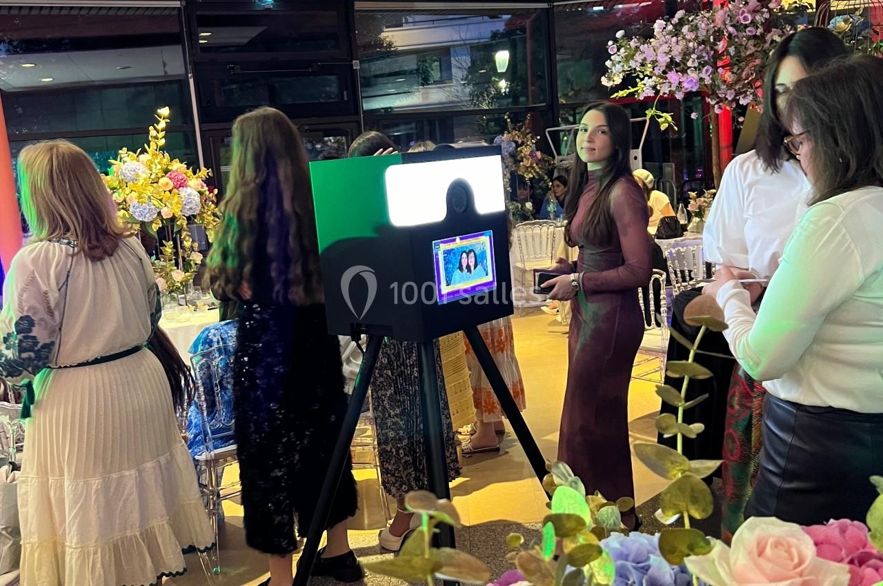 Des personnes se tiennent près d'une borne photo dans une salle décorée de fleurs et éclairée par des lumières colorées.