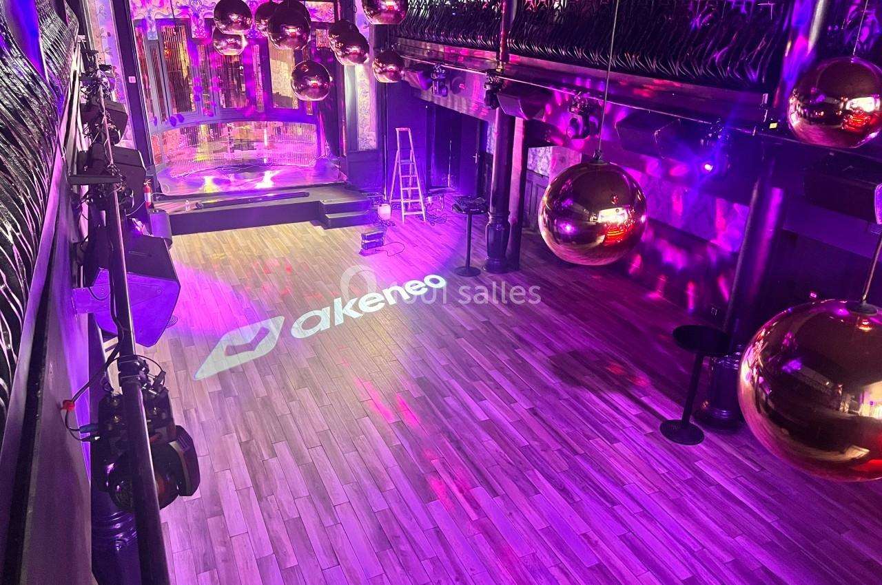 Salle de réception vide avec parquet, décor lumineux, boules dorées suspendues et logo projeté au sol.