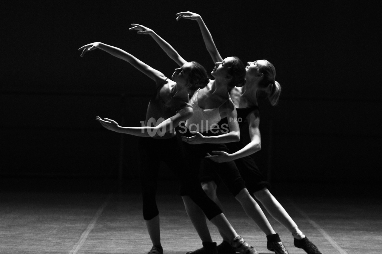 Trois danseuses exécutant une chorégraphie synchronisée sur une scène sombre, en noir et blanc.