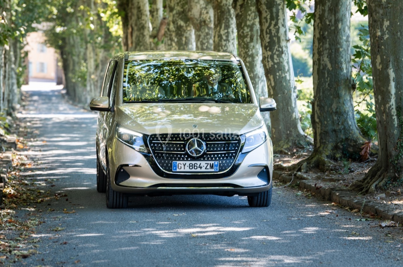 Monospace argenté Mercedes-Benz roulant sur une route bordée d'arbres, sous une lumière tamisée.