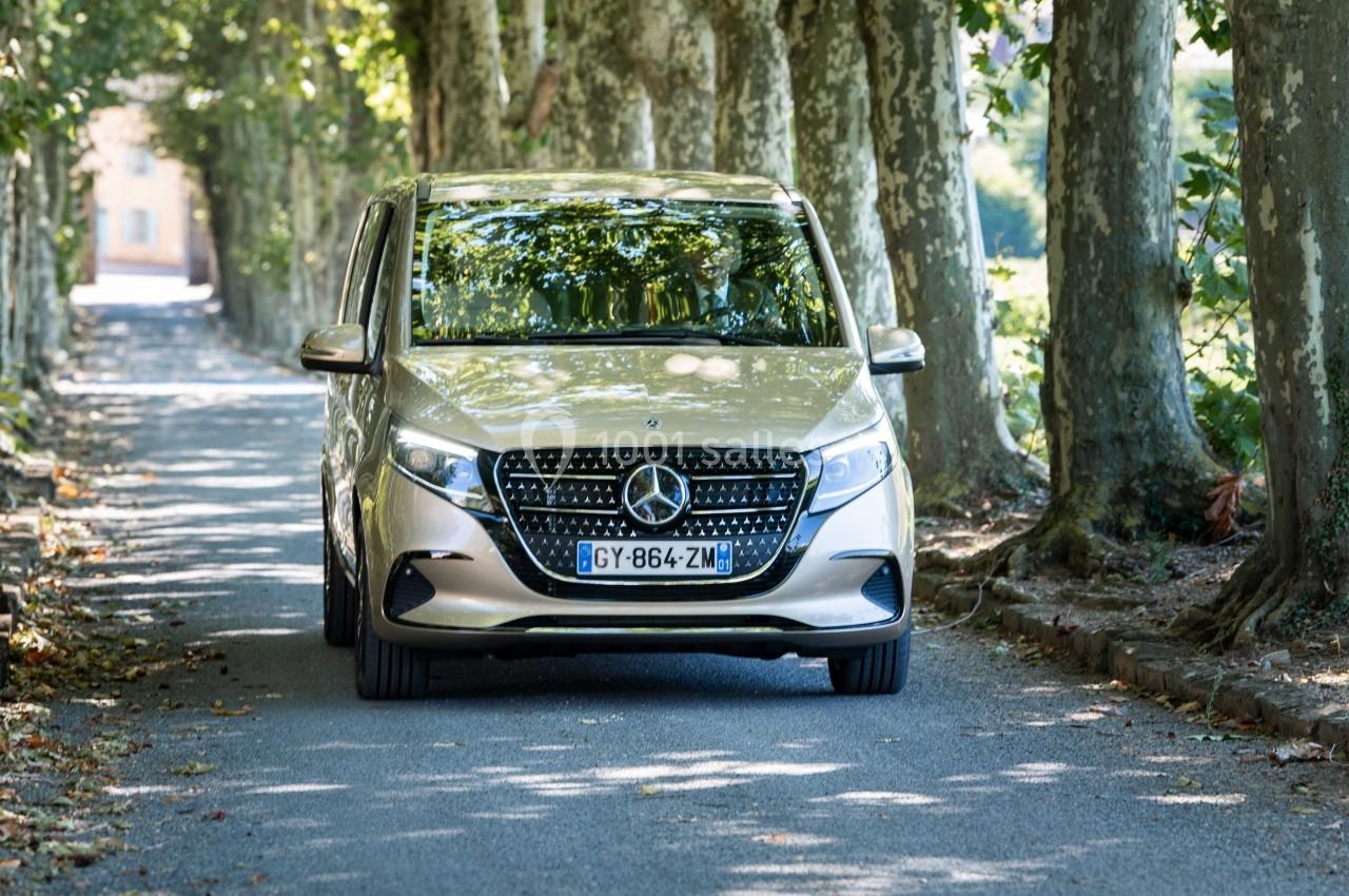 Monospace argenté Mercedes-Benz roulant sur une route bordée d'arbres, sous une lumière tamisée.