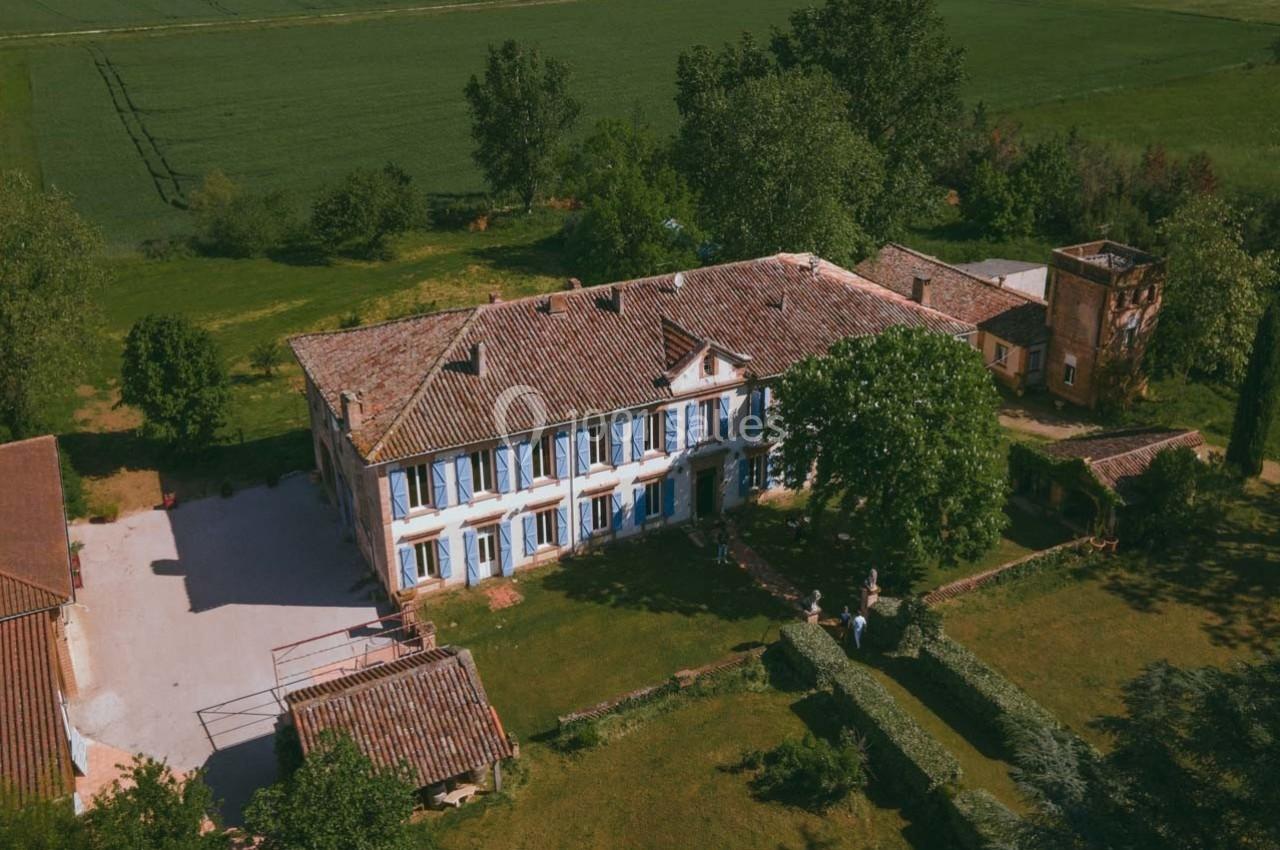 Vue aérienne d'une grande maison de campagne avec volets bleus, entourée de verdure et de champs.