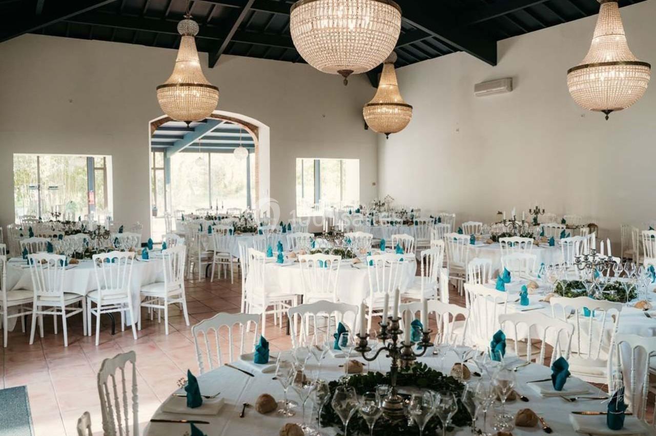 Salle de réception lumineuse avec tables rondes dressées, nappes blanches et serviettes bleues, décorée de lustres élégants.