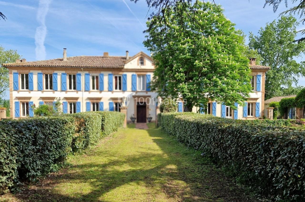 Grande maison de campagne avec volets bleus, entourée de haies et d'arbres, sous un ciel dégagé.