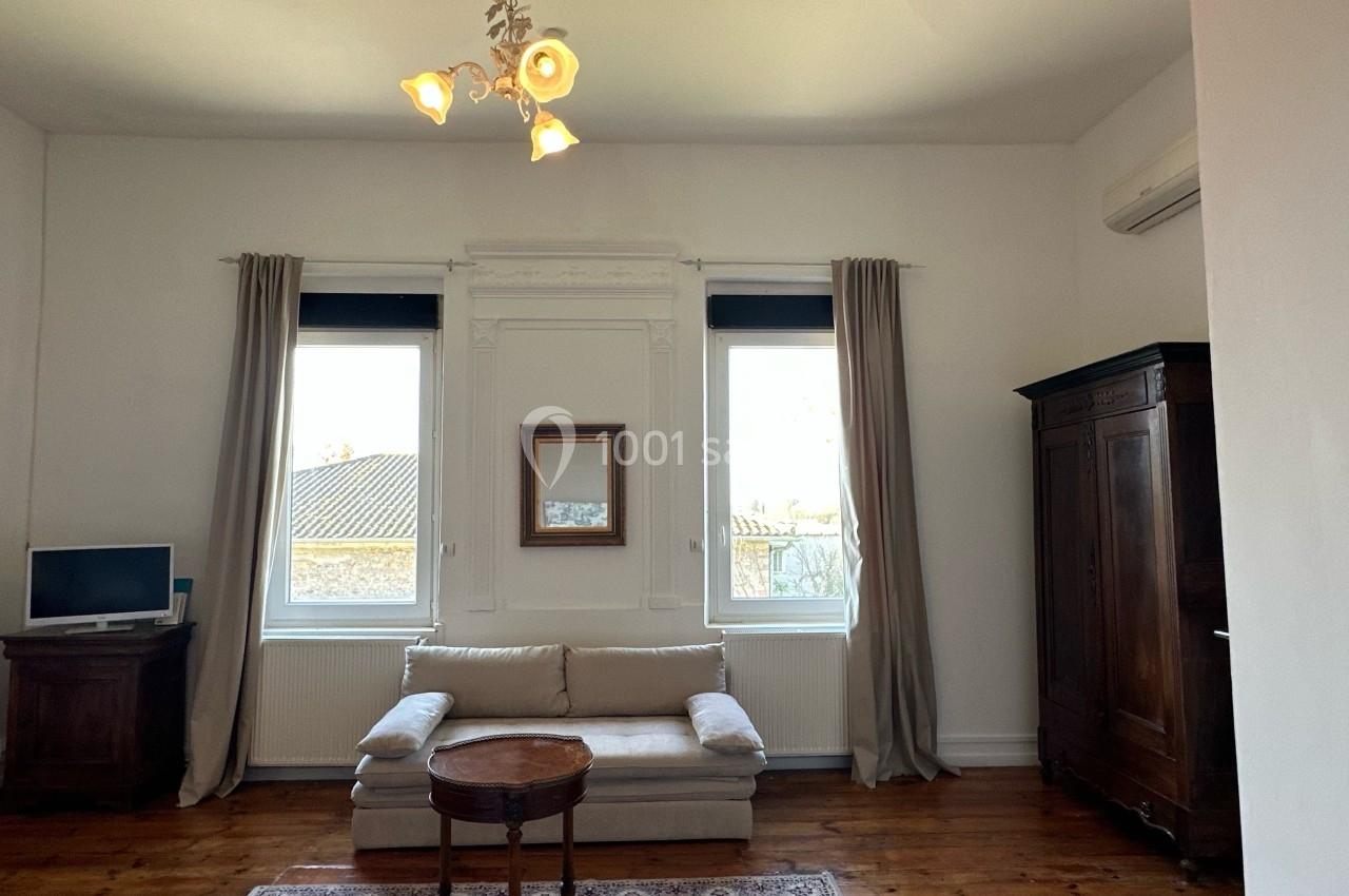 Salon lumineux avec canapé beige, table basse en bois, armoire ancienne, téléviseur et deux grandes fenêtres avec rideaux.
