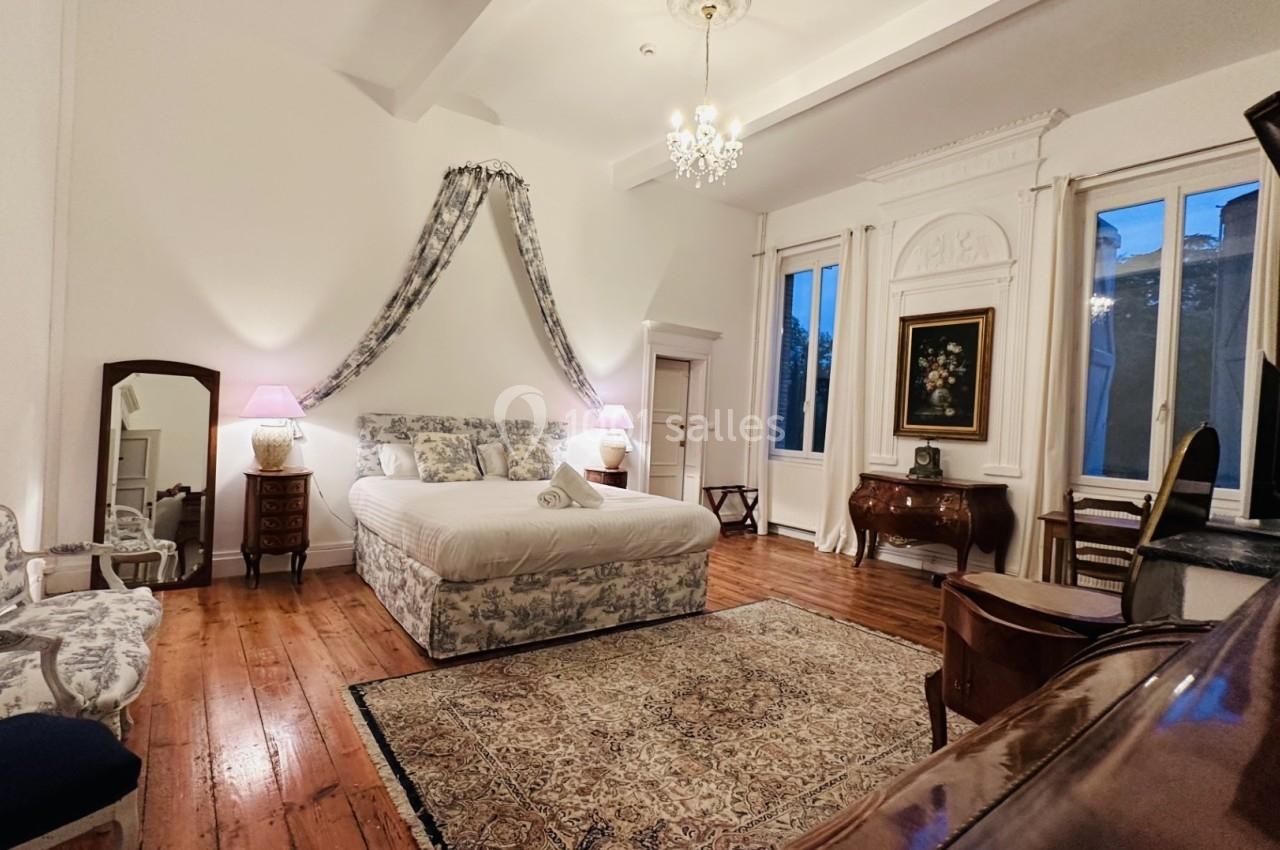 Chambre élégante avec lit double, mobilier ancien, tapis, et grandes fenêtres laissant entrer la lumière naturelle.