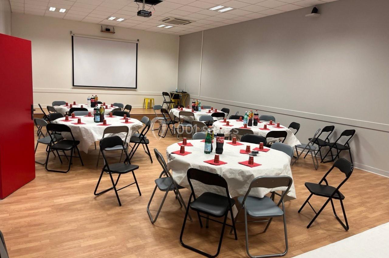 Salle aménagée avec des tables rondes dressées, nappes blanches, chaises pliantes noires et un écran de projection.