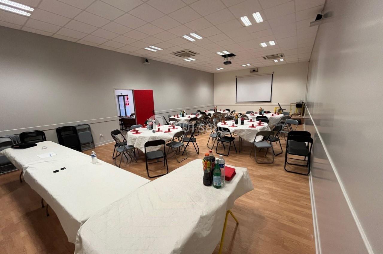 Salle de réunion avec tables disposées en U, chaises noires, nappes blanches et écran de projection au fond.