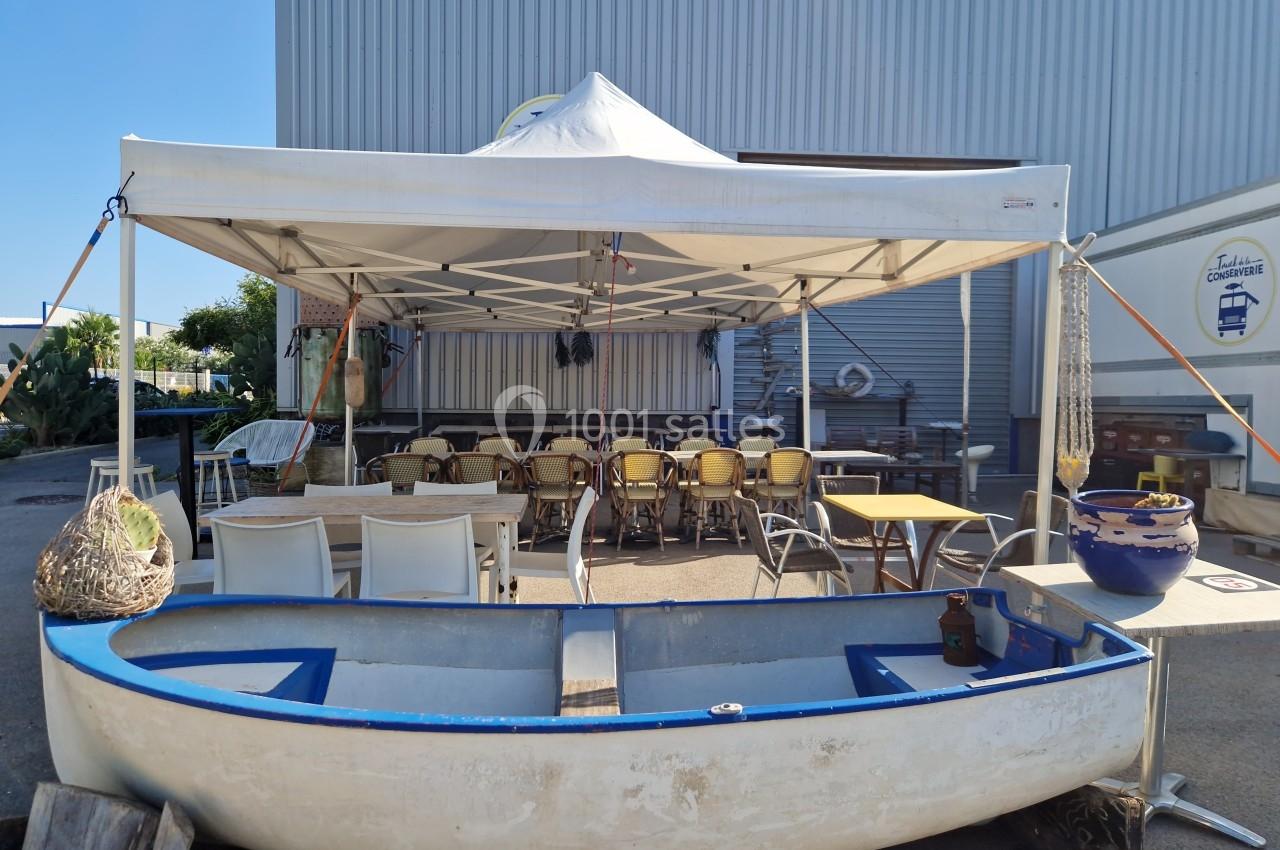 Une barque blanche devant une tente abritant des tables et des chaises, située près d'un bâtiment industriel.