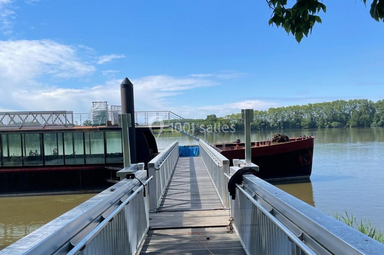 Passerelle en bois et métal menant à deux péniches amarrées sur une rivière, avec un pont visible à l'arrière-plan.