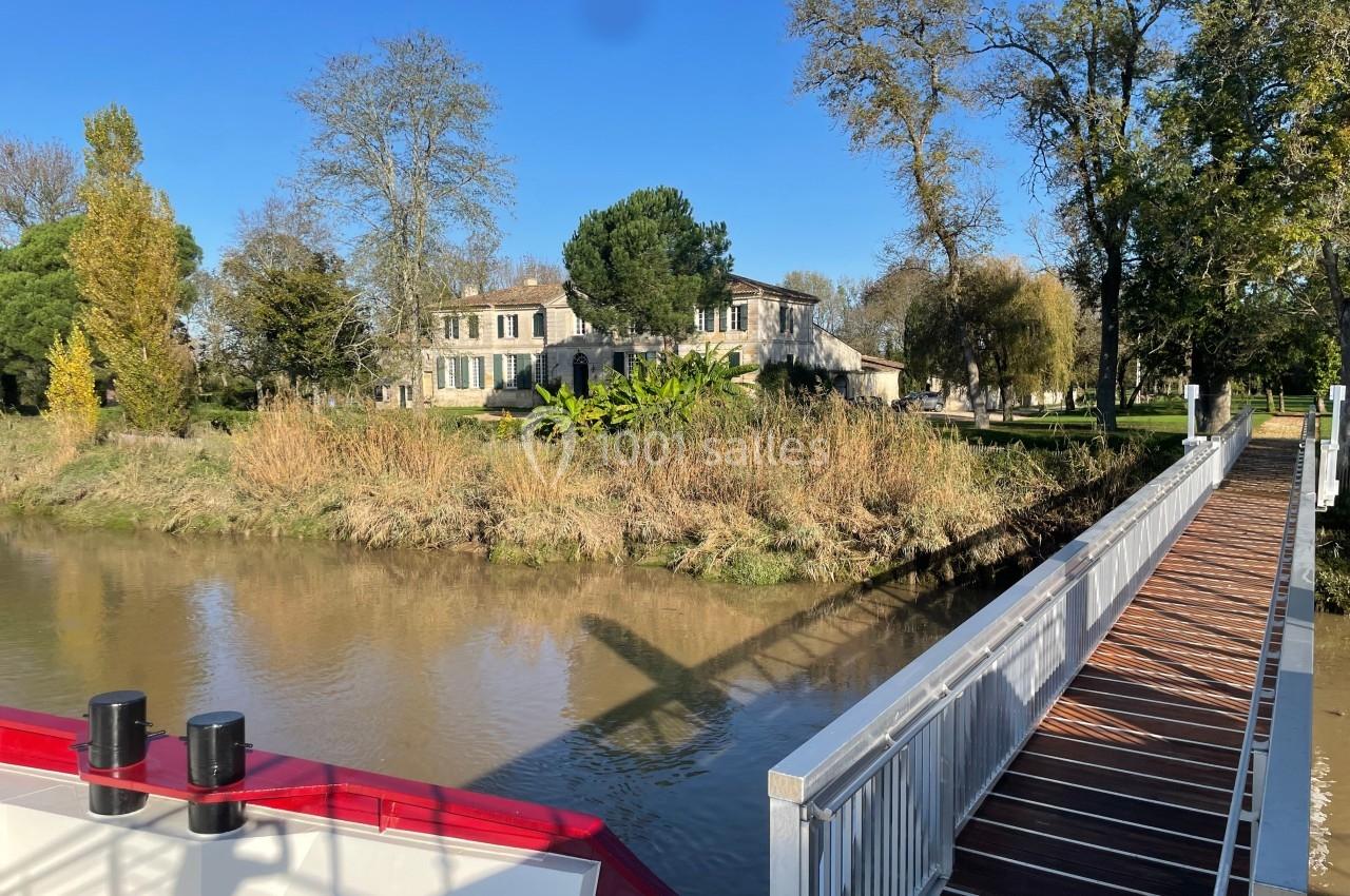 Maison en pierre entourée d'arbres, située au bord d'un canal avec une passerelle en bois et un bateau au premier plan.
