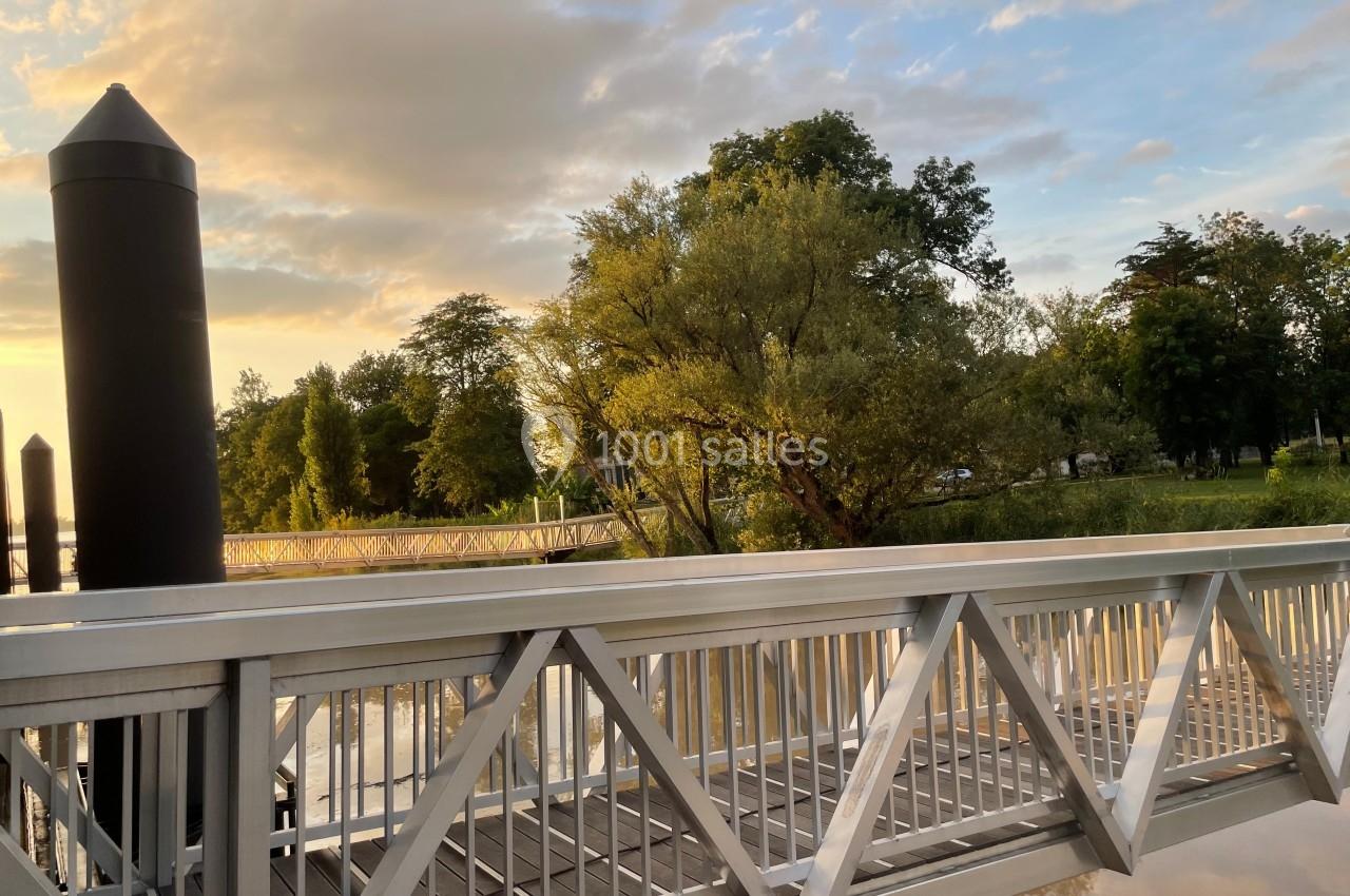 Passerelle métallique traversant une rivière bordée d'arbres, éclairée par une lumière douce au coucher du soleil.