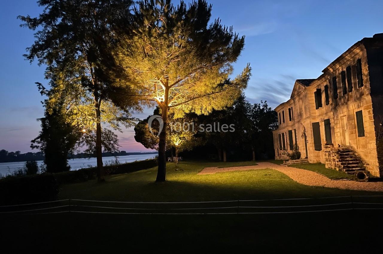 Façade d'une maison en pierre éclairée la nuit, entourée d'arbres et d'un jardin, avec une rivière en arrière-plan.