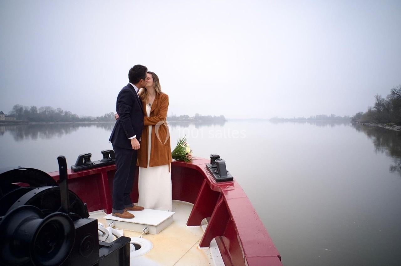 Un couple s'embrasse sur le pont d'un bateau, avec une rivière calme et un paysage brumeux en arrière-plan.