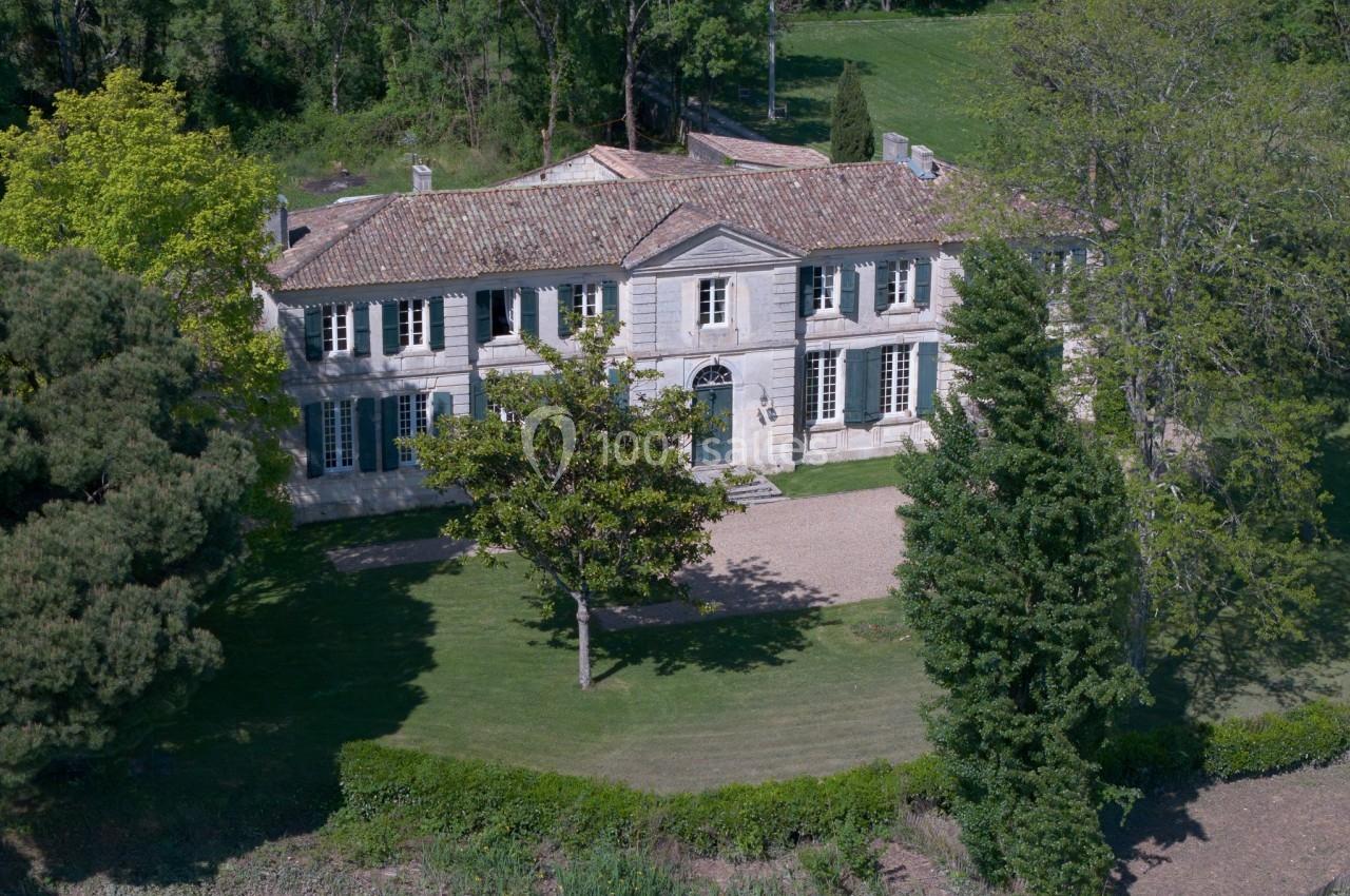 Grande maison en pierre à deux étages avec toit en tuiles, entourée de pelouse, arbres et végétation.