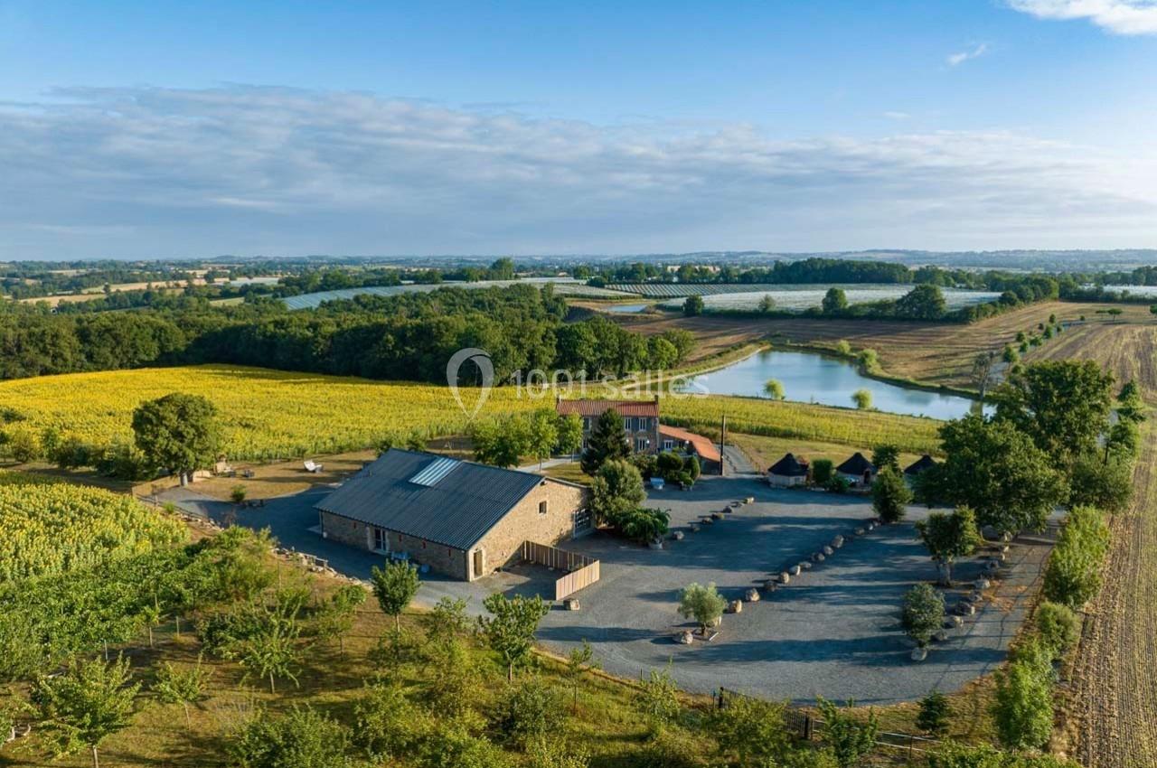 Vue aérienne d'une ferme en pierre entourée de champs, d'arbres et de plans d'eau sous un ciel partiellement nuageux.