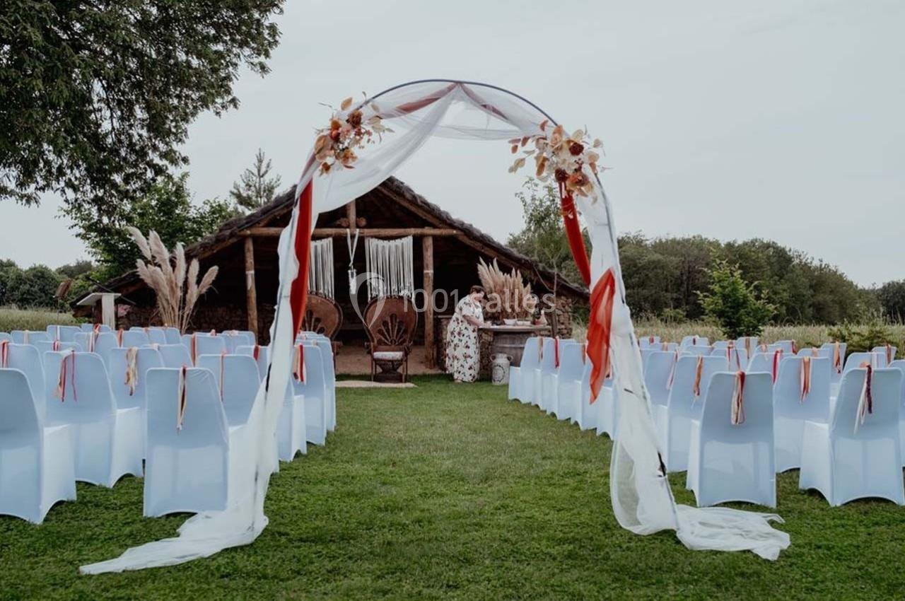 Allée centrale décorée pour une cérémonie en plein air, avec arche fleurie et chaises blanches sur pelouse.