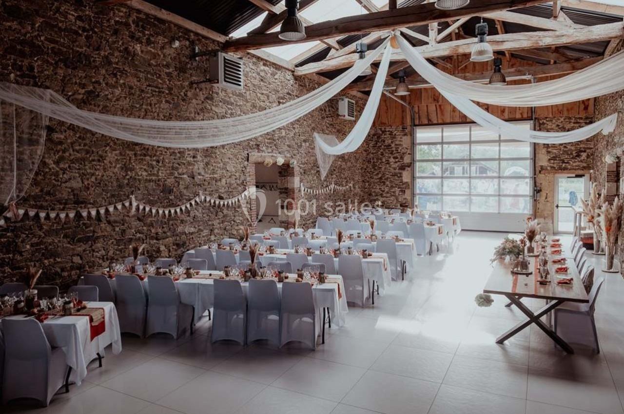 Salle de réception en pierre avec tables décorées pour un événement, éclairée par une lumière naturelle et des voilages…