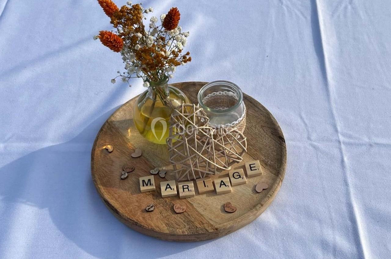 Centre de table avec vase de fleurs séchées, bougie, décoration en bois et lettres formant ’MARIAGE’ sur un plateau rond.