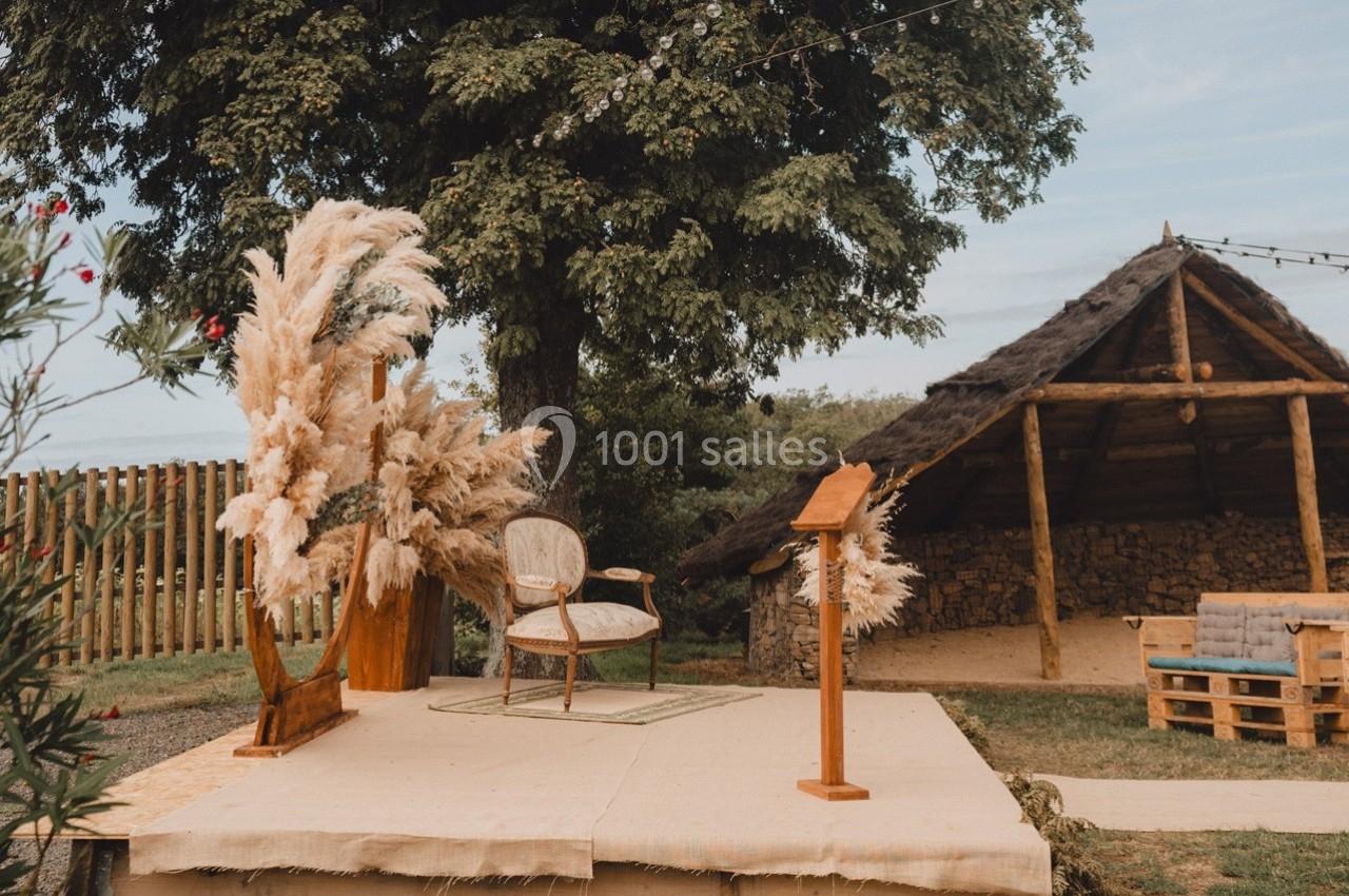 Estrade en bois décorée de pampas et d'un fauteuil, située en extérieur près d'une cabane rustique.