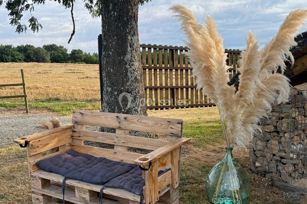 Banc en palettes avec coussins, vase en verre contenant des herbes de la pampa, posé dans un jardin champêtre.