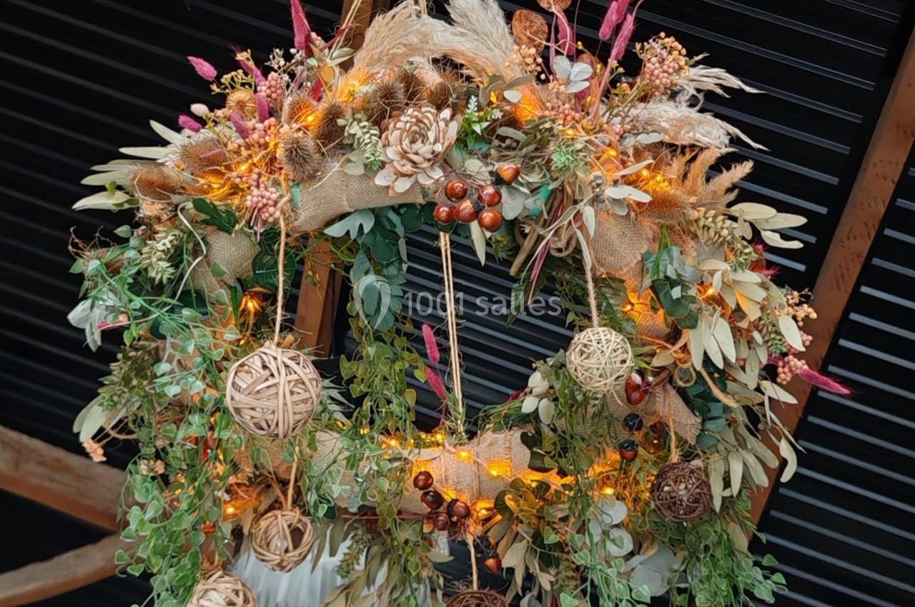 Couronne décorative en feuillage, pommes de pin, baies et lumières, suspendue devant un fond sombre en bois.