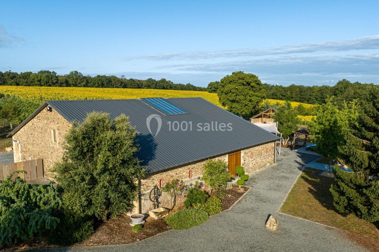 Bâtiment en pierre avec toit en tôle, entouré de végétation et de champs sous un ciel dégagé.