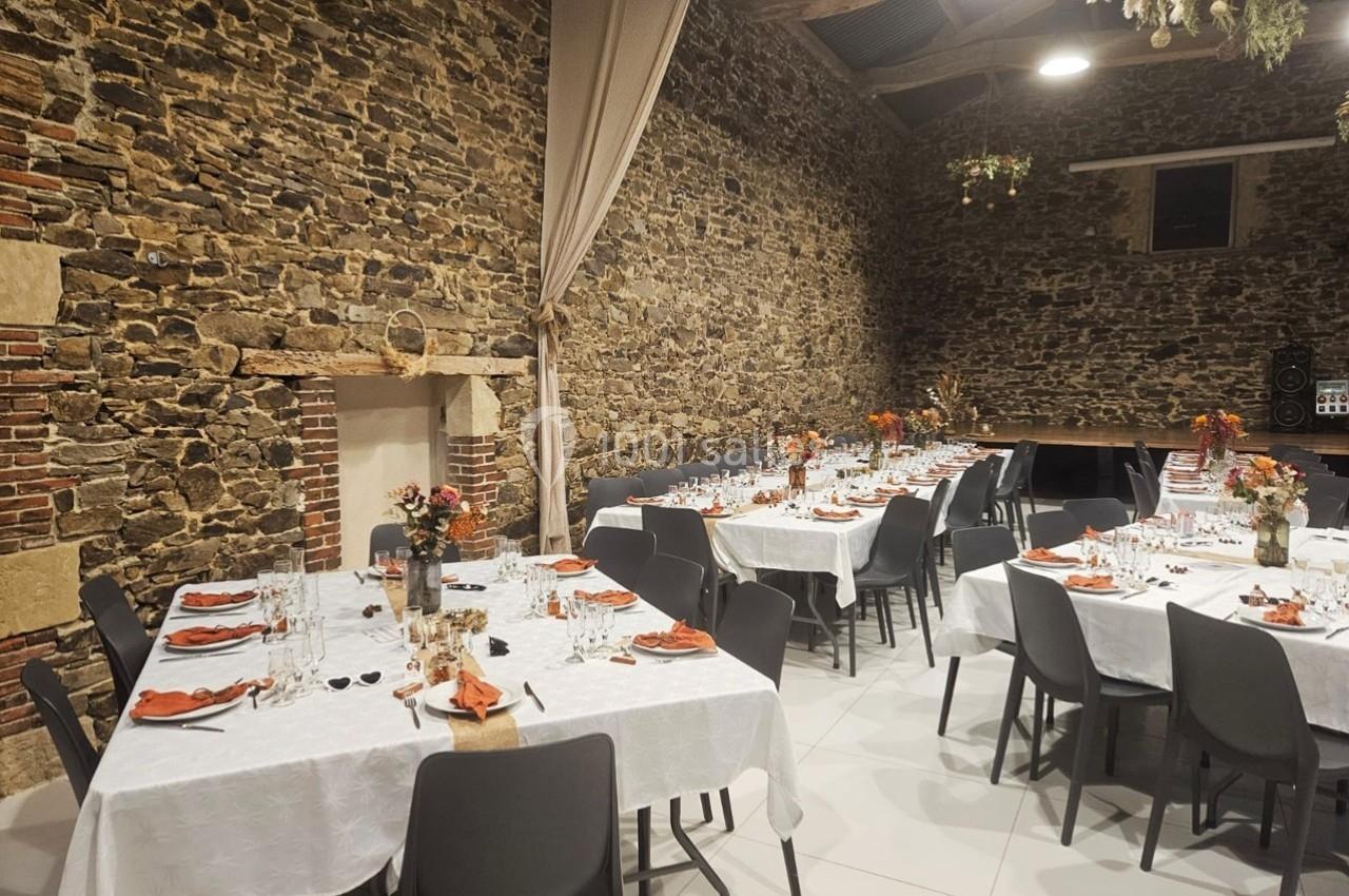 Salle en pierre aménagée avec des tables dressées pour un repas, décorées de fleurs et de serviettes orange.