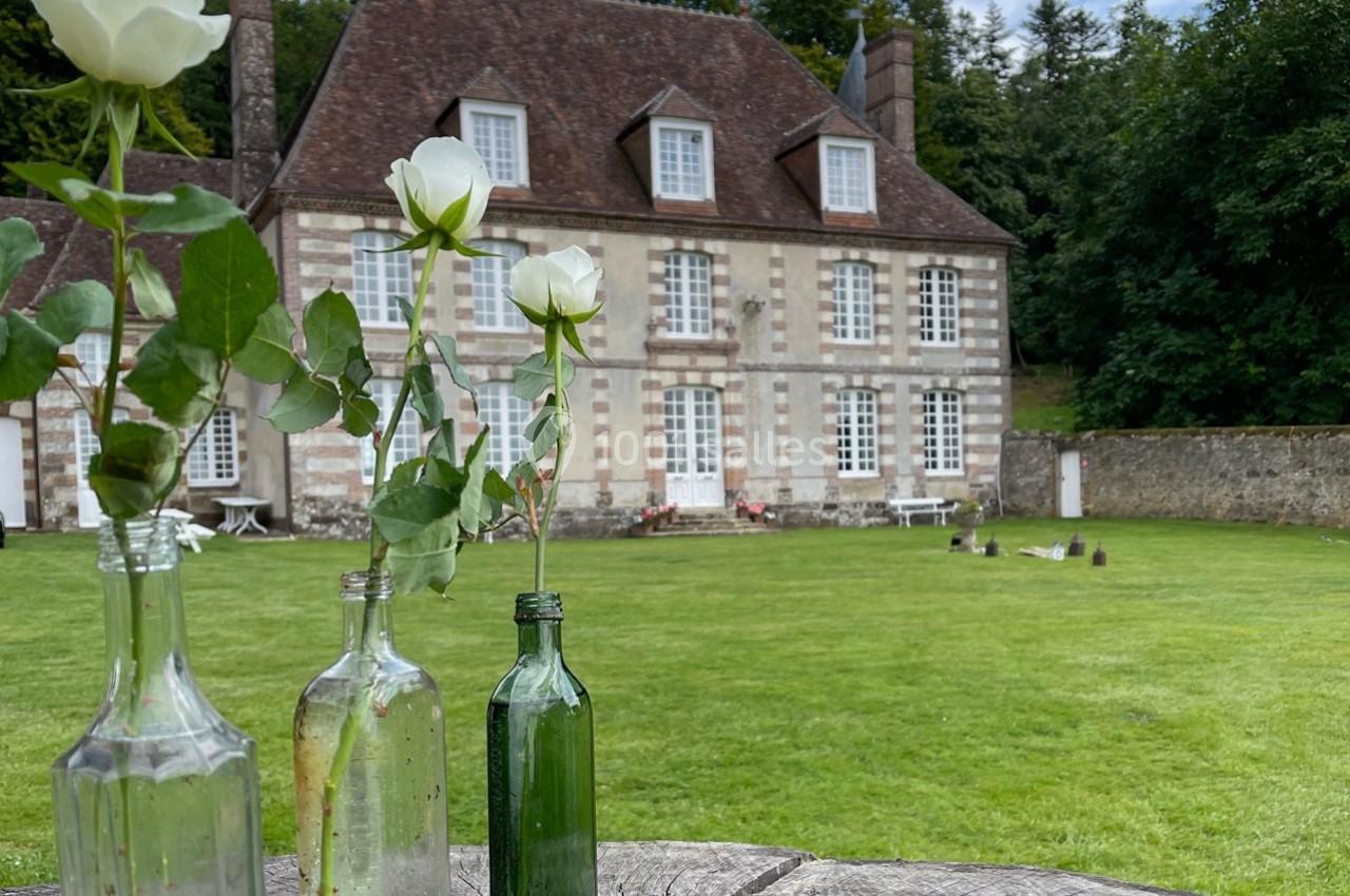 Roses blanches dans des bouteilles en verre sur une table en bois devant une maison en pierre avec un grand jardin.