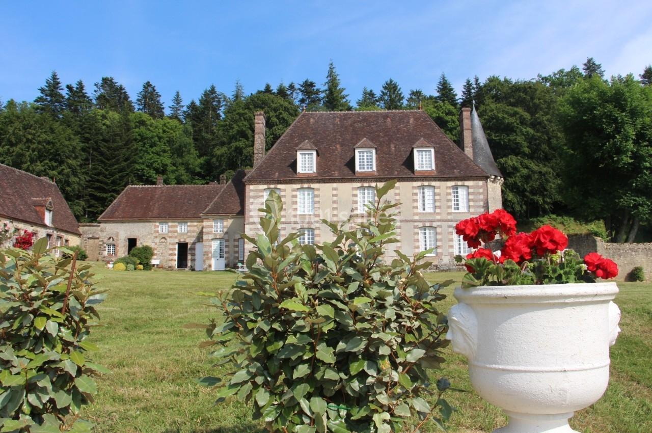 Manoir en pierre entouré de verdure, avec des rosiers et un grand jardin sous un ciel bleu.