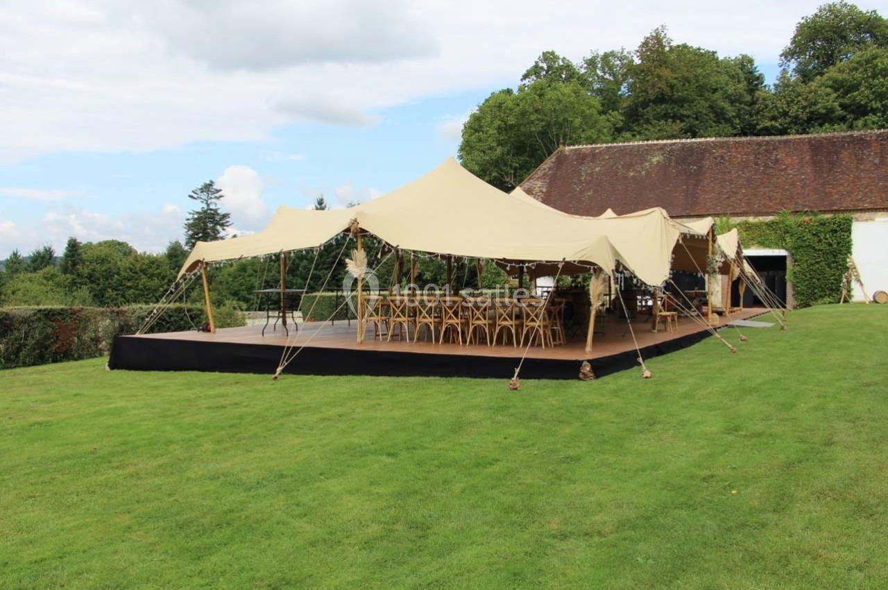 Tente beige montée sur une pelouse, équipée de tables et chaises en bois, entourée de verdure et d'un bâtiment en arrière…