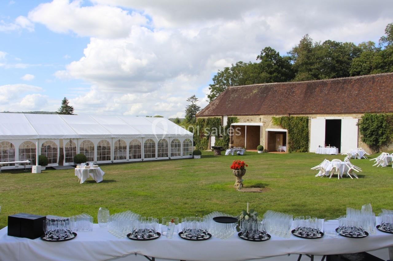 Grande pelouse avec une tente blanche, une grange en pierre et des tables dressées pour un événement en plein air.