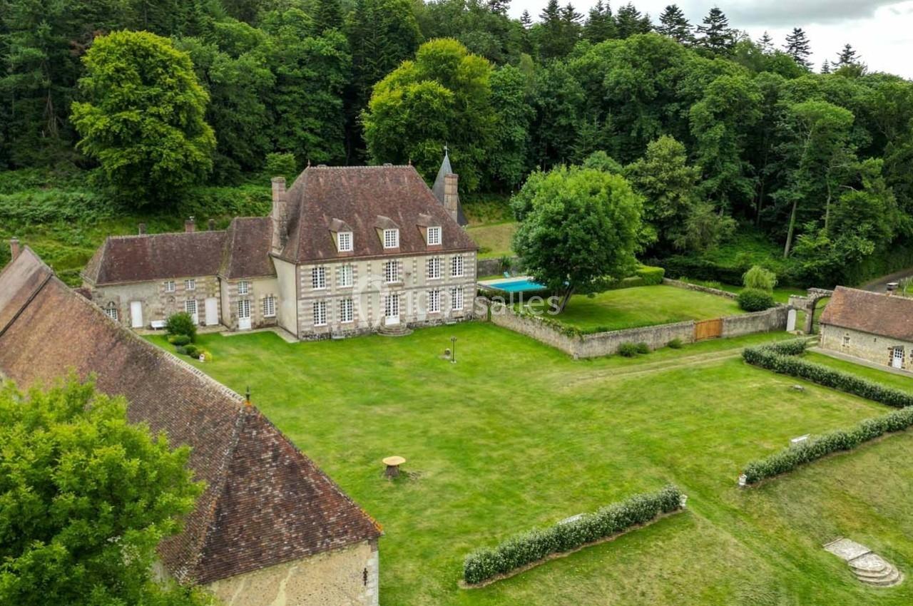 Vue aérienne d'un manoir entouré de bâtiments annexes, situé dans un grand jardin verdoyant bordé d'arbres.
