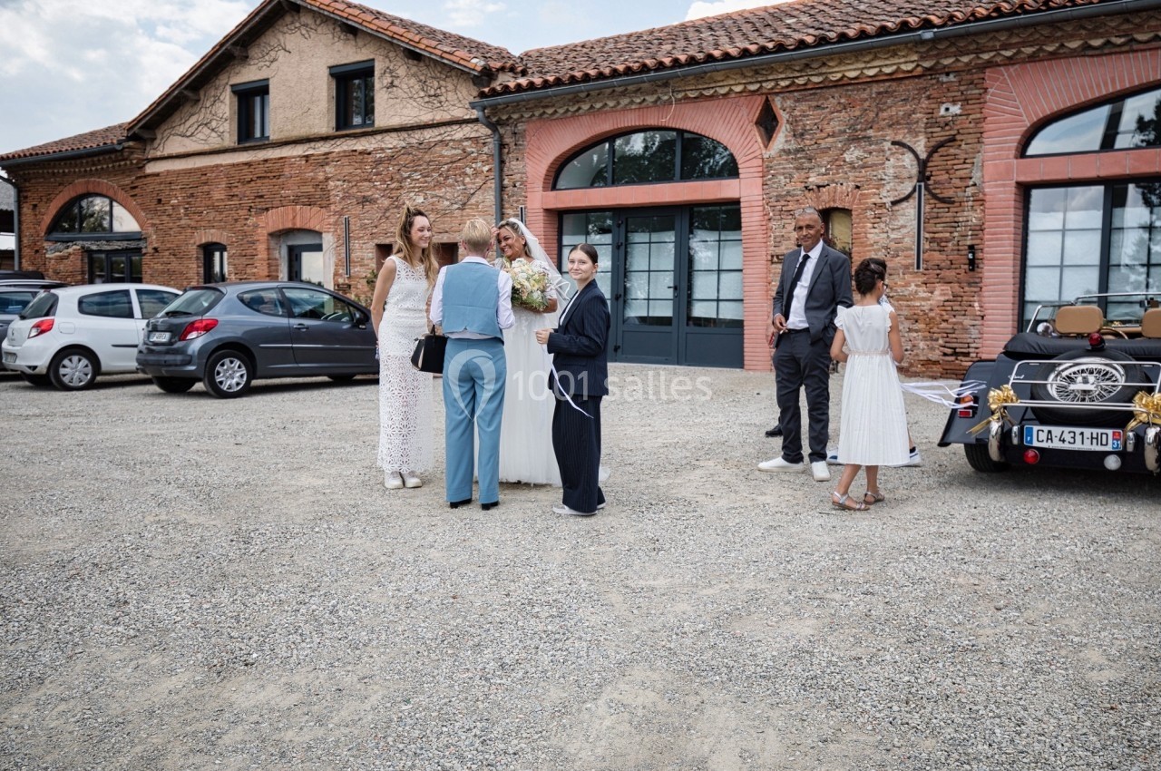 Un groupe de personnes discute devant un bâtiment en briques, avec une voiture ancienne stationnée à droite.