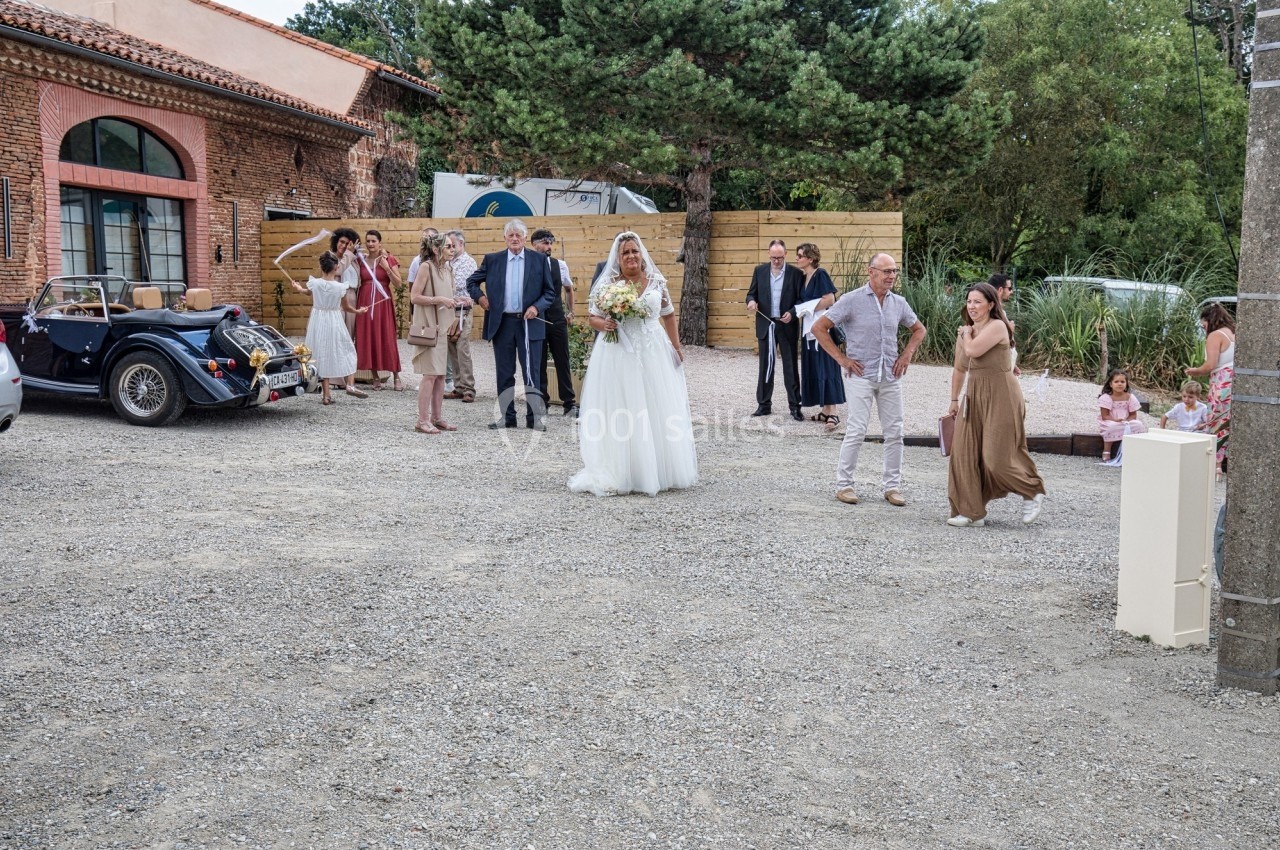 Une mariée en robe blanche marche vers un groupe de personnes dans une cour, avec une voiture ancienne en arrière-plan.