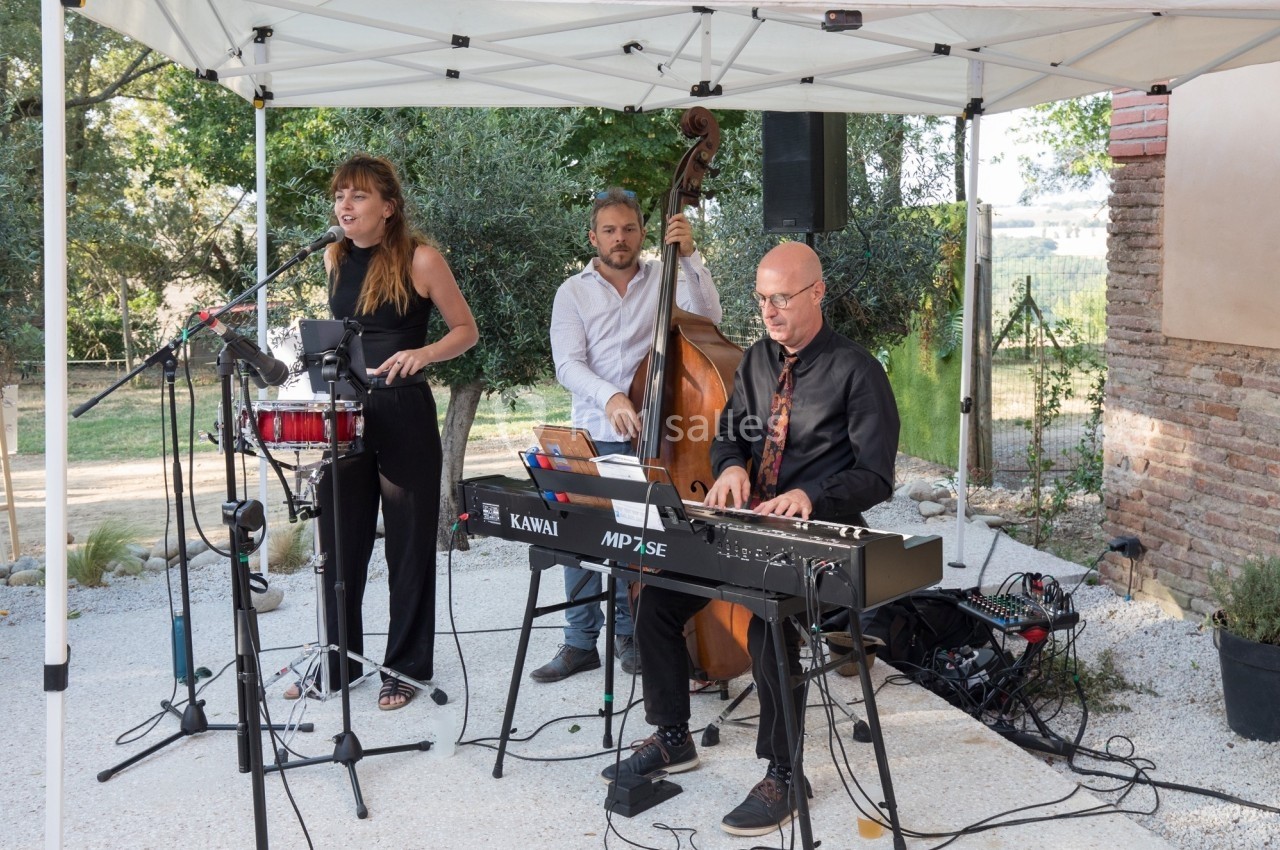 Un trio de musiciens joue en plein air sous une tente, avec une chanteuse, un contrebassiste et un pianiste.